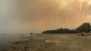 Photo taken on a beach showing trees and kayak’s on the right, the shore, and Lake Tahoe on the left and above it all is a dense cloud of smoke.
