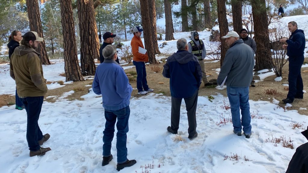 Un grupo de hombres y mujeres están de pie formando un círculo al aire libre en un paisaje boscoso escuchando hablar a un hombre.